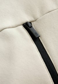 Tessuto beige con una superficie liscia, dotato di una cerniera nera e una piccola lettera 'B' in rilievo sul tirante della cerniera.