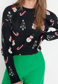 Czarny sweter z dzianiny z świątecznymi wzorami bałwanka, choinki i laski cukrowej; z okrągłym dekoltem, długimi rękawami, w połączeniu z zielonymi spodniami.