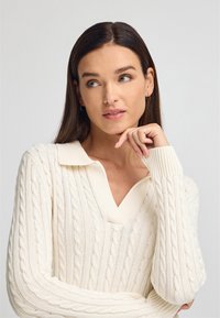 Strickpullover in cremiger Farbe mit Zopfmuster, langen Ärmeln und einem kragenlosen V-Ausschnitt-Design. Auffällige gerippte Bündchen und Saum.