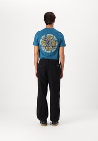 Turquoise short-sleeve t-shirt with circular graphic design featuring yellow roses and black accents, paired with loose-fitting black pants.