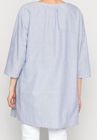 Tunika a righe azzurro chiaro e bianco con maniche a tre quarti, vestibilità comoda, piega sul retro e dettagli in bottone sulle maniche. Materiale in cotone.