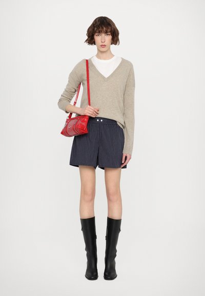 Un pull beige en V sur un t-shirt blanc, associé à un short à rayures verticales bleu marine. Un sac à main rouge et des bottes noires montantes complètent le look.