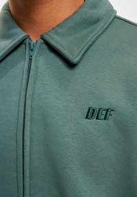 Kreklu ar rāvējslēdzēju, izgatavots mīksta auduma un ar struktūru, pelēkzaļā krāsā, ar izšūtu "DEF" logotipu kreisajā pusē.
