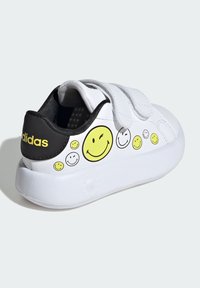 White leather sneakers with black heel, yellow smiley face designs, and Velcro straps. Textured black interior, solid white sole.
