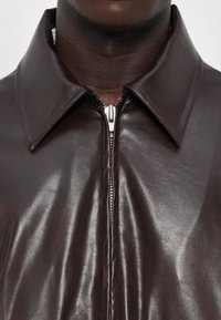 Smeđa kožna jakna s glatkom teksturom, sa oštrim okovratnikom i srebrnim patentnim zatvaračem sprijeda. Nema vidljivih uzoraka ili akcenta.