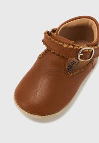 Καφέ δερμάτινο παπουτσάκι μωρού με κυματιστές άκρες και λουράκι με ασημένια πόρπη σε ανοιχτό φόντο.