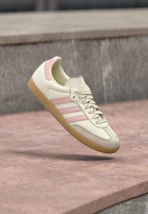 Zapatilla de cuero beige con detalles en rosa, que presenta tres franjas laterales, una puntera redondeada y una suela de goma texturizada. Cuello ligeramente acolchado.