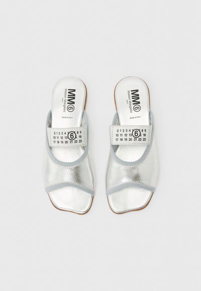 Ανοιχτά σανδάλια με ασημί χρώμα και ανοιχτά δάχτυλα, με γκρι περίγραμμα, φαρδιές τιράντες με αριθμούς και κείμενο "MM6 Maison Margiela Made in Italy" στα εσωτερικά πέλματα.