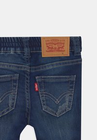 Jeans de mezclilla azul oscuro con una cintura elástica, que cuentan con dos bolsillos traseros y una etiqueta de cuero con marca que tiene costuras rojas y detalles de acento.