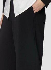 Pantalons larges noirs avec une texture lisse, dotés de poches latérales. Associés à une chemise blanche à boutons, mettant en avant un design épuré.