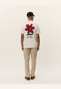 Camiseta blanca de algodón con un gran diseño de flor roja y hojas verdes, con el texto "Les Deux" en la parte inferior, llevada con pantalones beige.