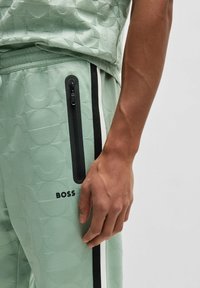 Ljusgröna joggers med geometrisk textur, med en sidficka med dragkedja, svarta sidostripes och en logga i svart text nära midjan.