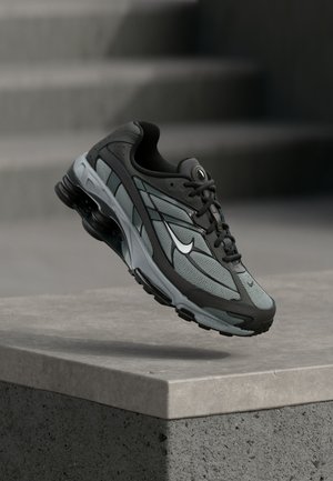 Pantof sportiv gri și negru cu panouri din plasă și talpă groasă, poziționat în aer deasupra unui prag de beton, cu scări pe fundal.