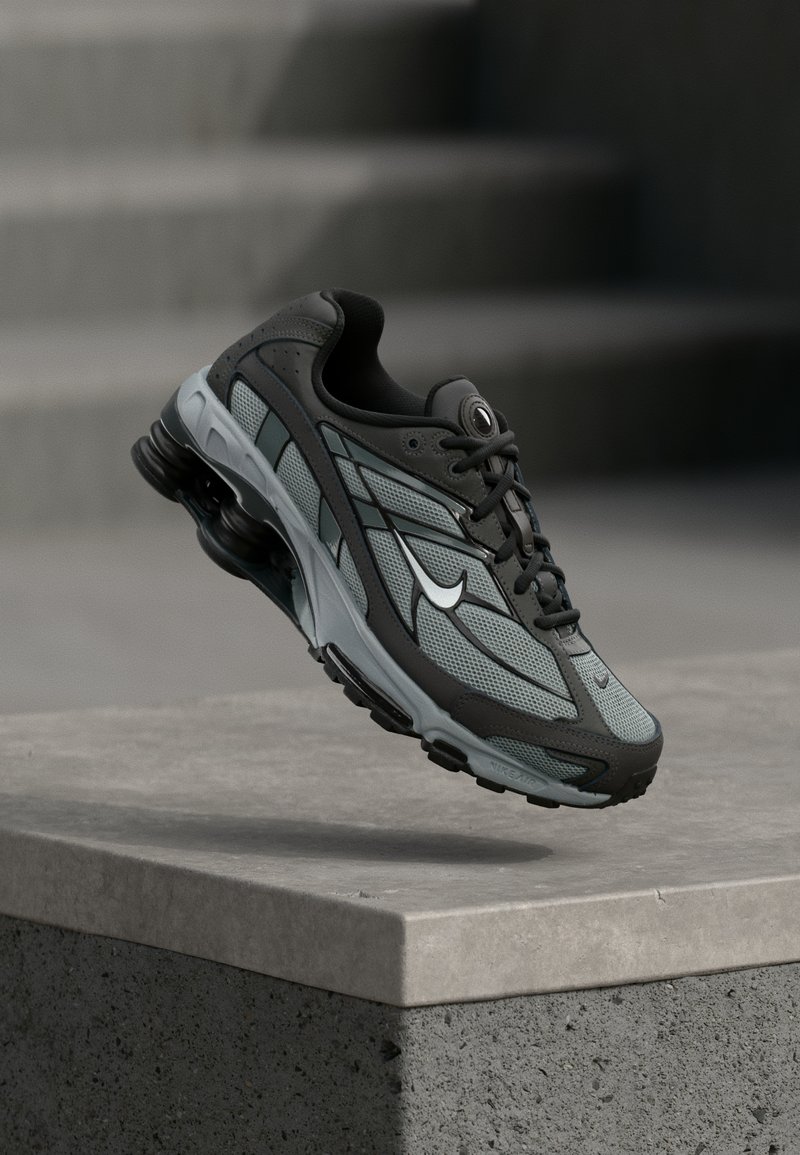 Γκρι και μαύρο αθλητικό παπούτσι με πλέγμα και χοντρή σόλα, τοποθετημένο στον αέρα πάνω από ένα τσιμεντένιο περιθώριο με σκάλες στο βάθος.