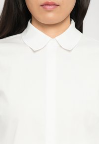 Ingrandimento di una persona che indossa una camicia bianca con colletto, con lunghi capelli scuri visibili su uno sfondo neutro.