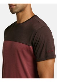 Camiseta de manga corta en marrón oscuro y burdeos, hecha de una tela suave. Presenta un diseño de bloques de color en contraste y un logo rojo en la manga.