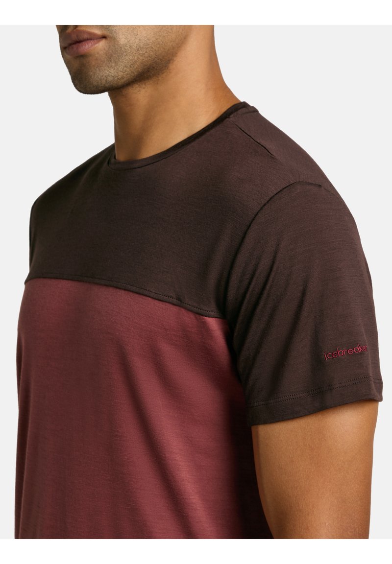 Camiseta de manga corta en marrón oscuro y burdeos, hecha de una tela suave. Presenta un diseño de bloques de color en contraste y un logo rojo en la manga.
