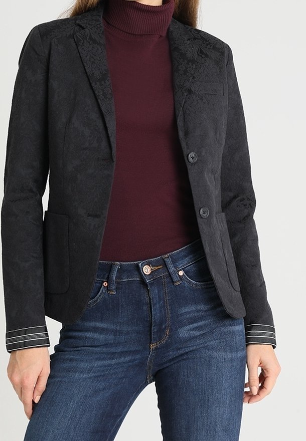 Femme portant un blazer ajusté à motif floral noir sur un col roulé bordeaux et un jean bleu, sur un fond uni.