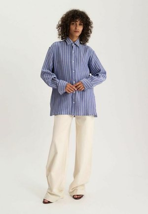 Femme aux cheveux bouclés porte une chemise à rayures verticales bleues et blanches, un pantalon large beige et des talons ouverts noirs, debout devant un fond uni.