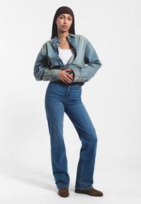 Chaqueta de mezclilla de corte relajado en azul claro, llevada sobre una camiseta sin mangas blanca. Vaqueros de tiro alto azul con corte recto y botas de ante marrón.