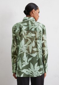 Chemise à manches longues en tissu transparent vert, ornée d'un motif botanique avec des feuillages de couleur vert clair et des poignets boutonnés.