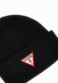 Berretto di maglia nera con una texture a coste. Presenta un patch triangolare rosso e bianco con testo nero che recita "GUESS USA".