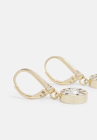 Des boucles d'oreilles en métal doré présentent un pendentif en cristal rond avec des accents gravés, fixé par un fermoir à clip. Fini métallique lisse.