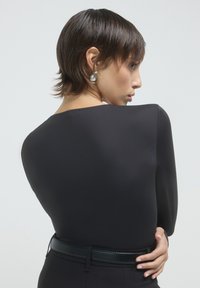 Top nero a maniche lunghe con una texture liscia e colletto a girocollo. La modella lo indossa con una cintura nera e orecchini a cerchio d'argento, visto da dietro.