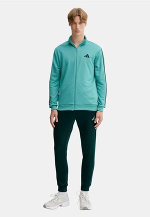 Giovane uomo con giacca teal con cerniera, pantaloni sportivi neri con loghi e scarpe da ginnastica bianche, in piedi contro uno sfondo chiaro e semplice.