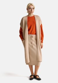 Krémový pletený dlouhý cardigan přes oranžový svetr a odpovídající krémovou sukni s žebrovanými detaily. Černé baleríny dotvářejí vzhled.