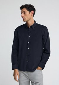Camicia blu navy con colletto, realizzata in tessuto morbido, con bottoni bianchi e un piccolo logo sul petto. Abbinata a jeans grigi.