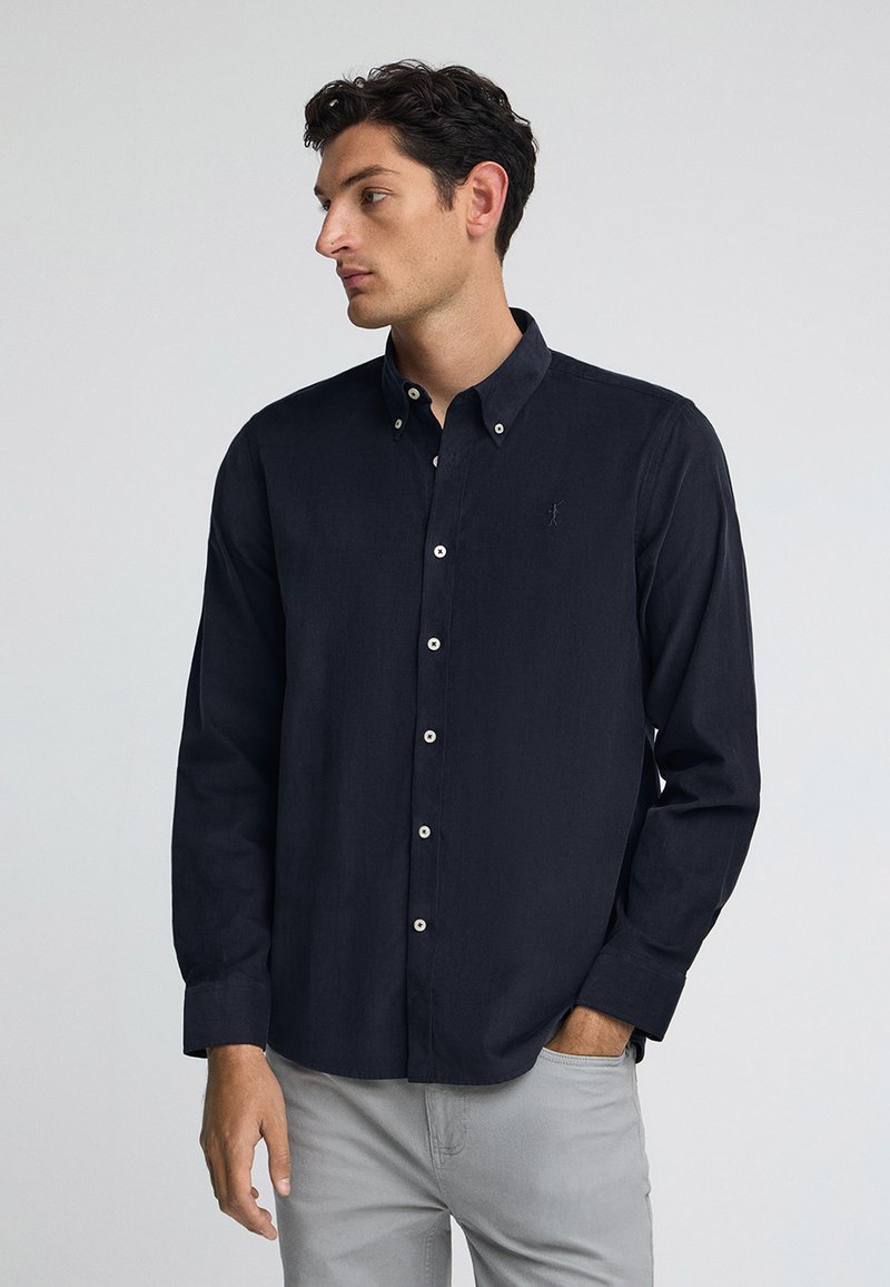Camicia blu navy con colletto, realizzata in tessuto morbido, con bottoni bianchi e un piccolo logo sul petto. Abbinata a jeans grigi.