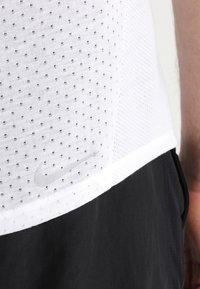Biały, perforowany podkoszulek z subtelnym logo Nike w odcieniu szarości, zestawiony z czarnymi szortami. Materiał ma gładką fakturę i przewiewny fason.