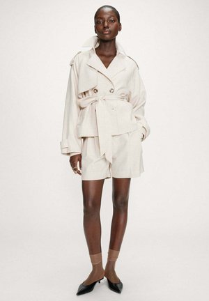 Femme portant un trench-coat crème ceinturé avec un short assorti, des chaussettes transparentes, des escarpins noirs à bouts pointus, et plusieurs bagues à la main gauche.