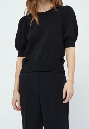 Femme portant un haut en maille noir à manches courtes bouffantes et un pantalon noir ample, les mains détendues le long du corps, sur un fond clair uni.