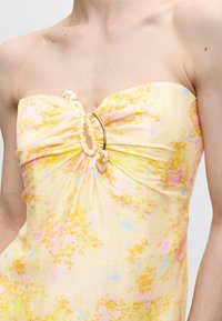 Κίτρινο floral φόρεμα χωρίς τιράντες με ρυτιδωμένη μέση και λεπτομέρεια με χρυσό κρίκο, φορεμένο σε γυμνούς ώμους και πάνω μέρος στήθους.