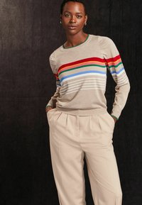 Pull en tricot beige avec des rayures horizontales colorées en rouge, vert, bleu et blanc ; associé à un pantalon crème plissé. Tissu texturé.