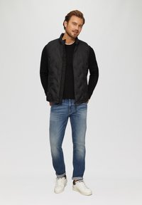 Zwarte, gewatteerde vest over een zwarte trui, gecombineerd met lichtblauwe jeans en witte sneakers, met een minimalistisch ontwerp en casual pasvorm.