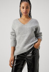 Grauer, gestrickter Pullover mit V-Ausschnitt, Rippstrickstruktur und lockerer Passform. Kombiniert mit glänzenden schwarzen Hosen, die eine glatte Oberfläche und einen taillierten Stil zeigen.