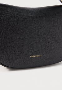 Borsa a mano in pelle nera granulata con forma curva e piccolo logo dorato "COCCINELLE" su sfondo bianco.
