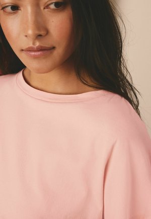 Nærbillede af en kvinde med langt mørkt hår iført en lyserød t-shirt med rund hals, der kigger let mod sin højre side mod en ensfarvet baggrund.