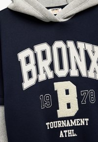 Marinblå hoodie med grå insida, med "BRONX" i vita bokstäver och en texturerad "B"-detalj. Sportinspirerad design med året 1978.