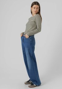 Gestreiftes Langarmoberteil in Beige und Schwarz, kombiniert mit blauen Jeans im weit geschnittenen Stil. Das Model trägt außerdem schwarze und graue Sneaker. Einfarbiger Hintergrund.