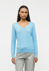 Világoskék, V nyakú pulóver, puha anyagból készült. Hosszú ujjakkal és bordázott szegéllyel rendelkezik. Világosszürke, laza szabású nadrággal párosítva.