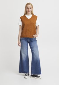 Orangen gestrickter Pullunder mit V-Ausschnitt und Strukturmuster, getragen über einem weißen Hemd. Kombiniert mit weit geschnittenen blauen Jeans und schwarzen Sneakers.