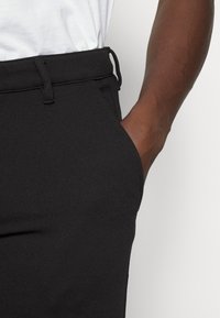 Svarta texturerade byxor med en midja som har en dragsko, med händer i fickorna, delvis visade mot en ljusgrå bakgrund.