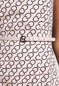 Robe sans manches avec un fond rose présentant un motif circulaire noir et blanc, accentuée par une fine ceinture rose avec une boucle en métal circulaire.