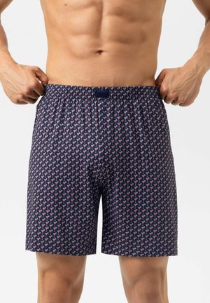 Homme portant un caleçon bleu marine orné de petits verres à cocktail roses, tenant la ceinture avec les deux mains.