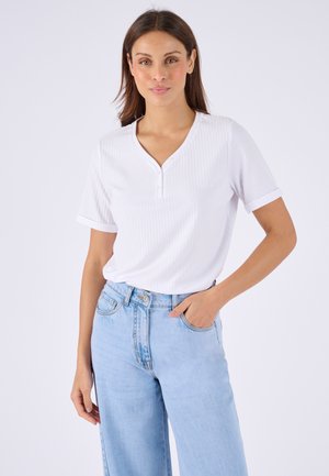 Donna con capelli castani lisci che indossa una maglietta henley bianca a maniche corte e jeans a vita alta azzurri, posando con una mano in tasca.