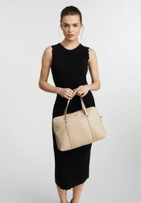 Beżowa skórzana torba na ramię z dwoma uchwytami, zamknięciem na zamek i pionowymi detalami szycia. Modelka ma na sobie czarną sukienkę bez rękawów z falbaniastym dołem.